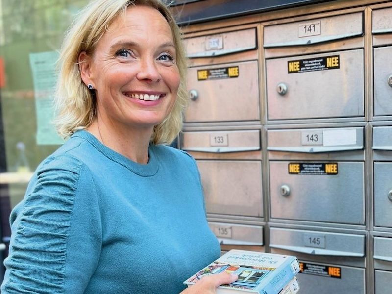 een dame met blond haar in een blauw shirt lacht en heeft in een haar handen een paar boeken van de bibliotheek ze staat voor een aantal brievenbussen