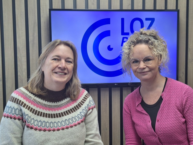 Twee dames zitten voor een beeldscherm met logo erop.