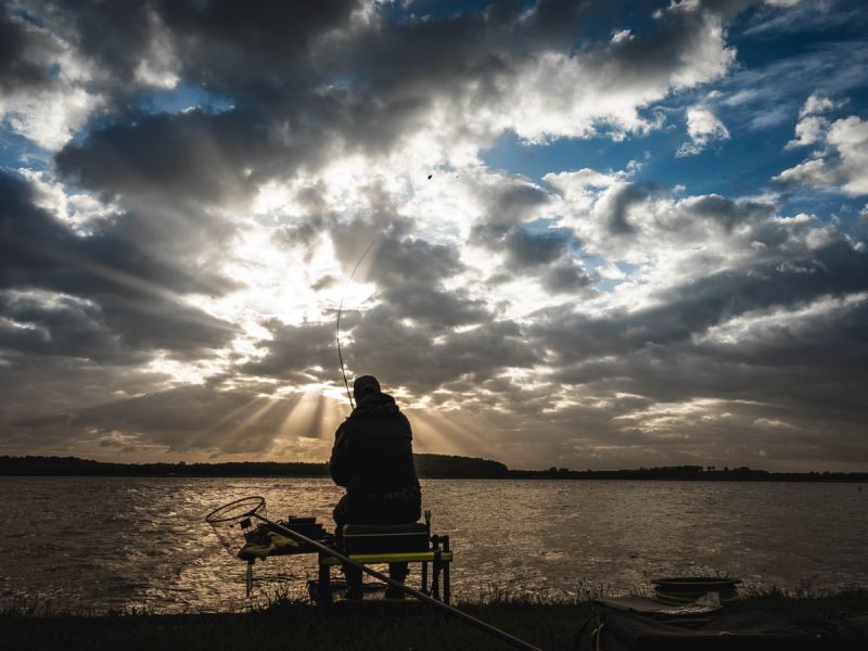 Een foto van een Visser bij het water met ondergaande of opkomende zon die door de wolken heen straalt