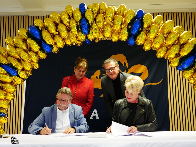 Twee mensen zitten aan tafel en tekenen een overeenkomst, twee mensen staan achter hen.
