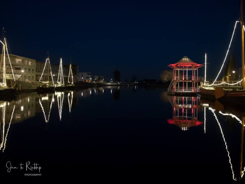Foto in het donker met bootverlichting en oranje huisje bij het water.