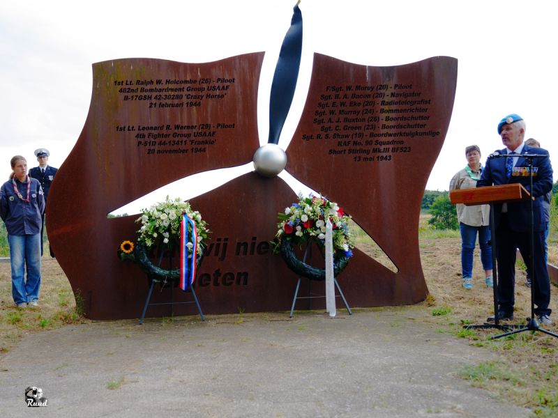 Aat Klop met 2 kransen bij het Vliegermonument, een Monument waar een vliegtuigpropellor in is te zien en de tekst opdat wij niet vergeten, daaarachter staan scouts