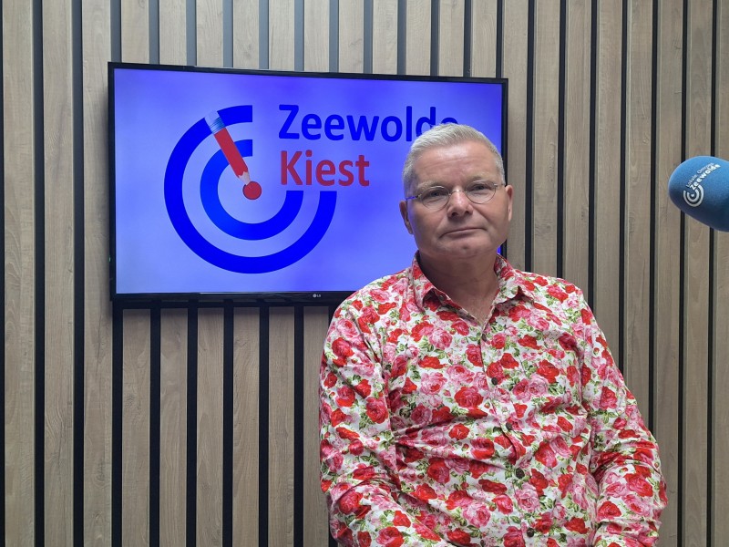 Man met bloemenshirt zit in studio voor een tv met logo erop.