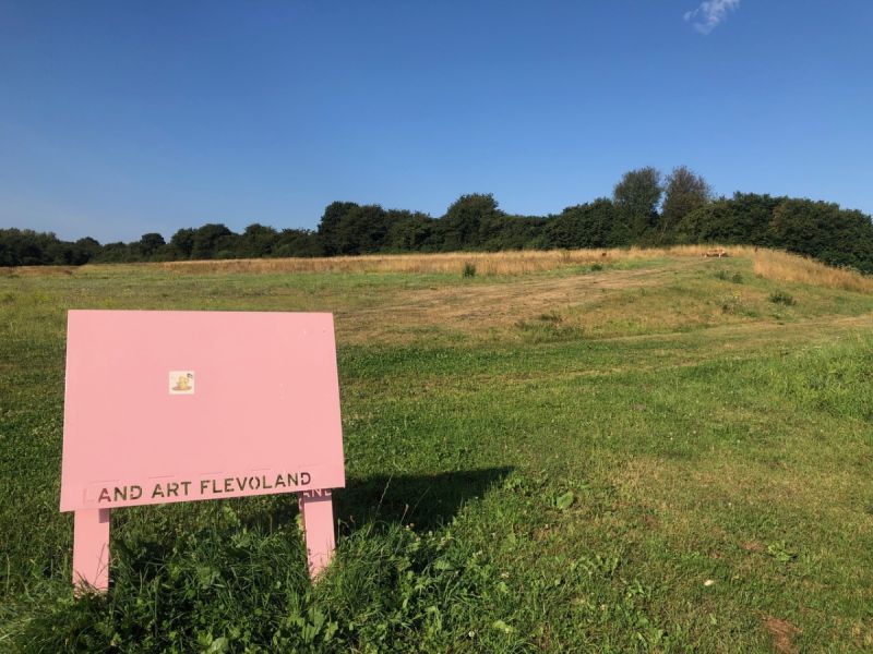 Roze bord van Land Art Flevoland met de Winnie the Pooh sticker met op de achtergrond SeaLevel verstopt achter hoog gras