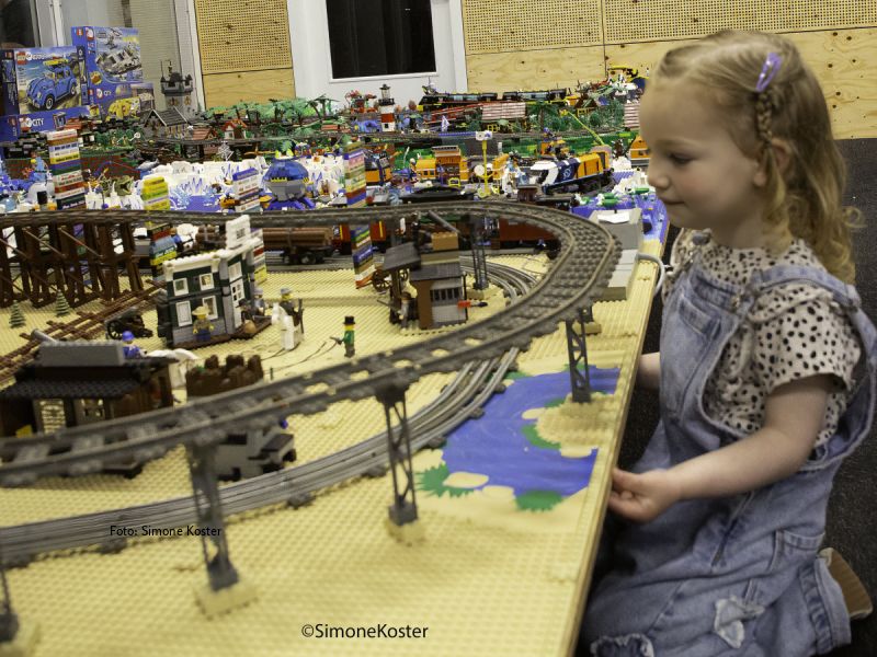 Een meisje kijkend bij een opstelling van rails waar trein overheen komt met op achtergrond nog meer lego