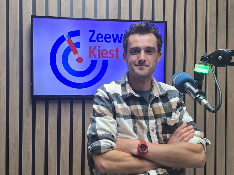 Man met rood horloge zit achter microfoon.