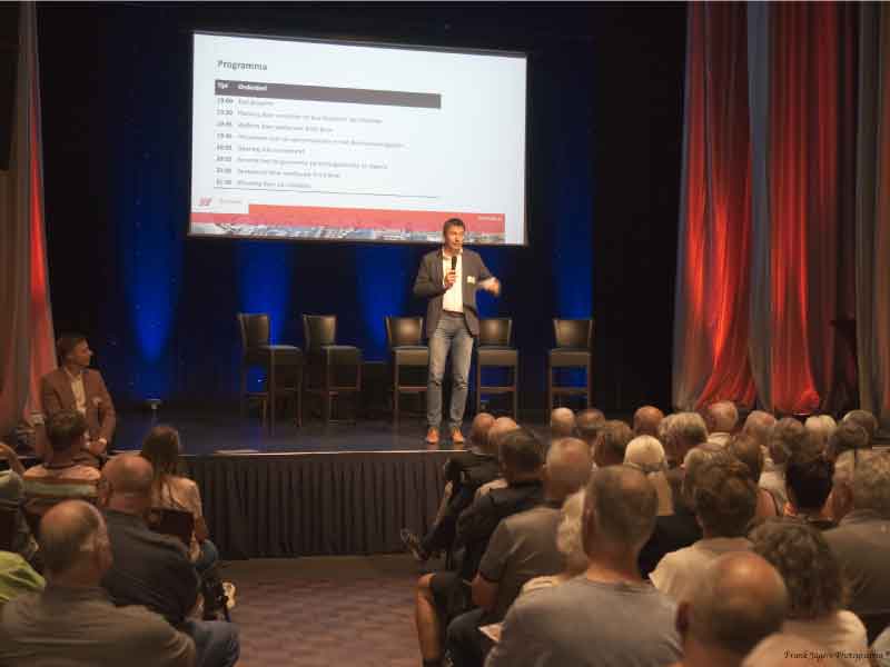 Een volle zaal in the lux met wethouder Bron op het podium en de startdia achter hem