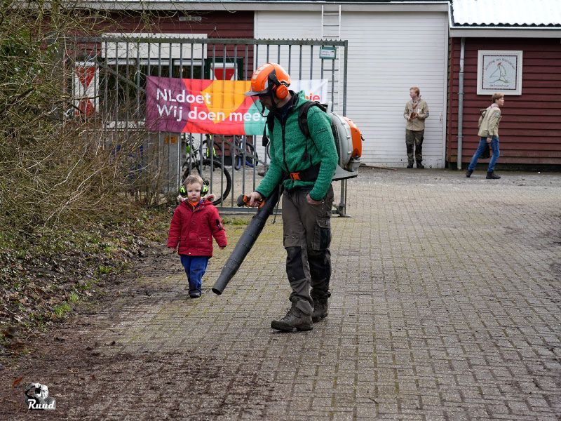 Een man met bladblazer voor het hek van scouting jan wandelaar, er loopt een klein kindje met hem mee