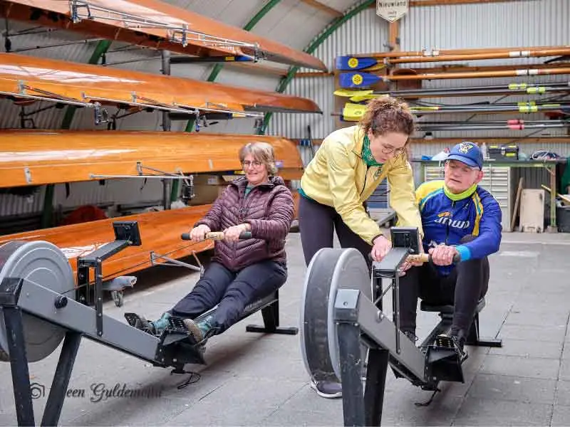 In de hal staan twee roeiapparaten, welkebeoefend worden door dame en heer, een trainster is er bij