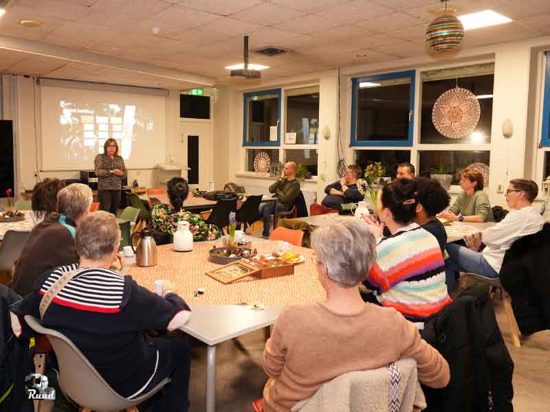Een groep vrijwilligers verdeeld over tafels in een zaal van de Meermin met Sanne als spreker voor een scherm
