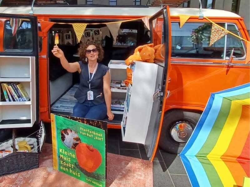 Een dame in de Biebbus, een vrolijke oranje bus met ervoor een kleedje en parasol, De vrouw heeft een ijsje in haar hand.