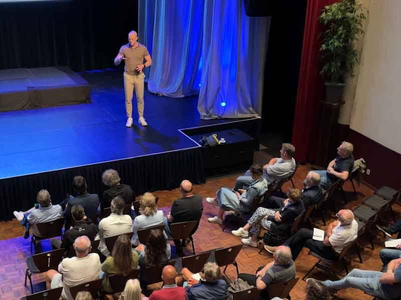 Presentator op het podium van de zaal in The Lux, publiek zit in een theateropstelling te luisteren