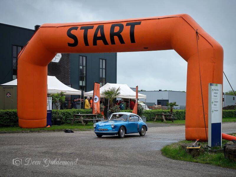 Grote oranje boog met het woord start eronder rijdt een klassieke blauwe auto