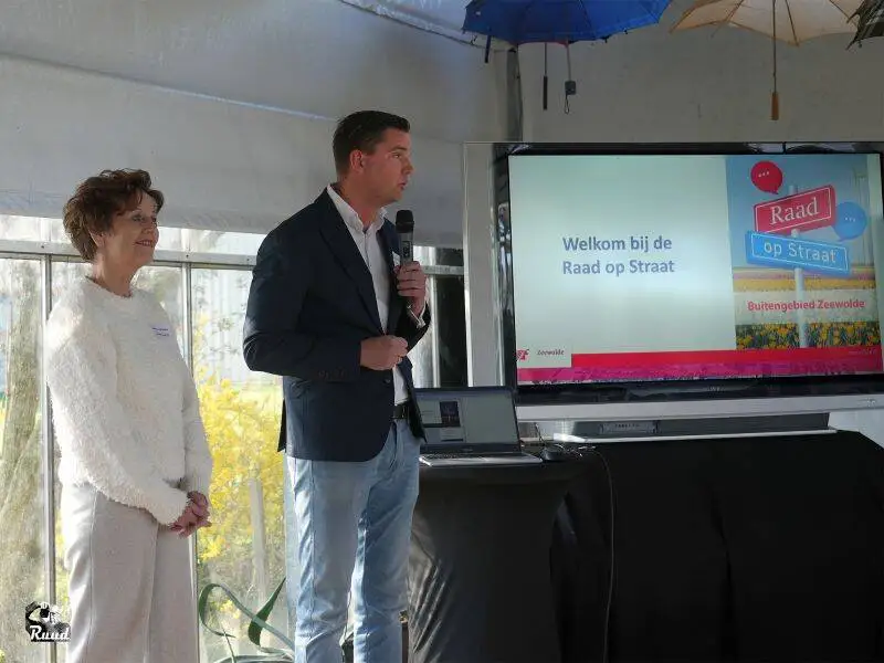 Twee mensen waarvan een met microfoon staan voor een scherm