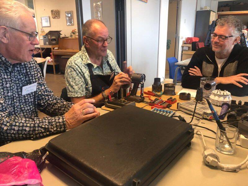 drie mensen zitten aan een tafel met gereedschap en spullen