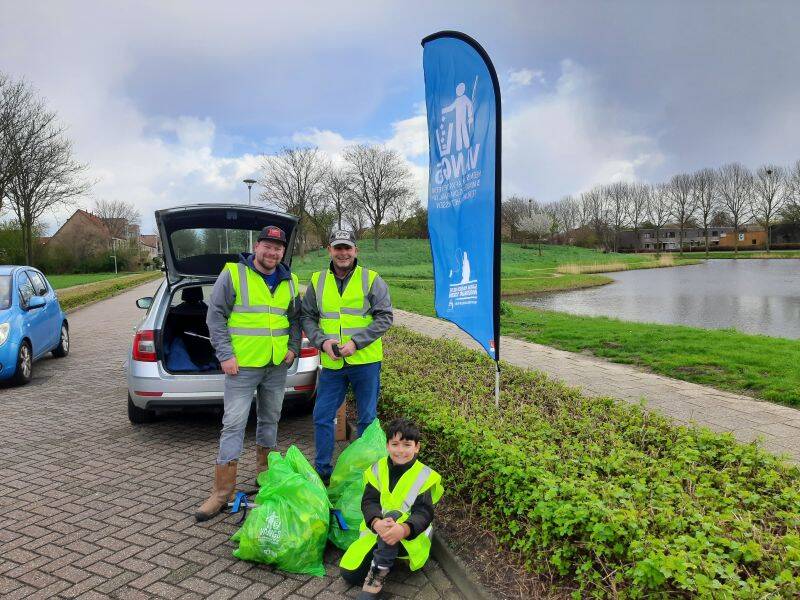 drie mensen met gele hesjes en groene afvalzakken staan bij het water naast een blauwe vlag