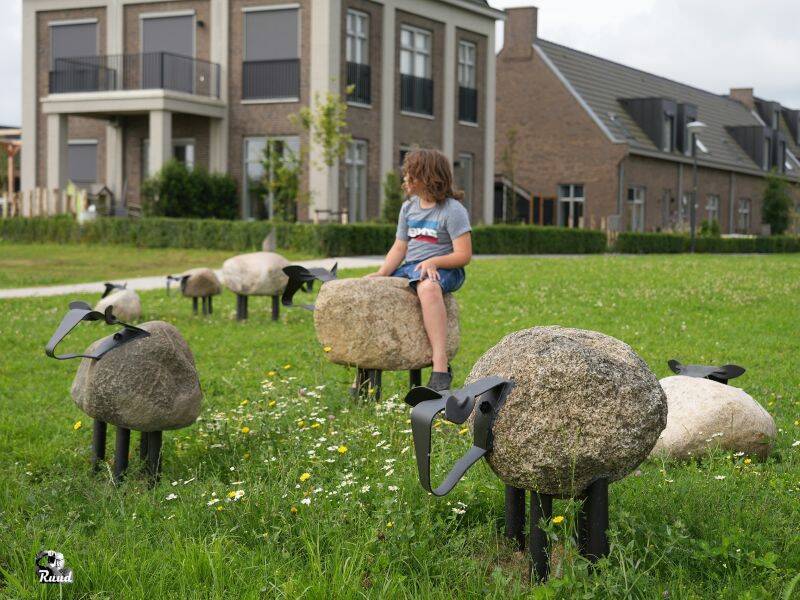 Stenen schapen in een groen grasveld met een huizen op de achtergrond en een jongen op een van de schapen.